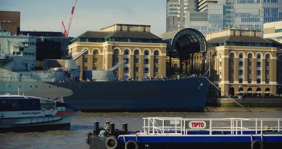 Ferry passes warship docked near buildings with construction crane in the background