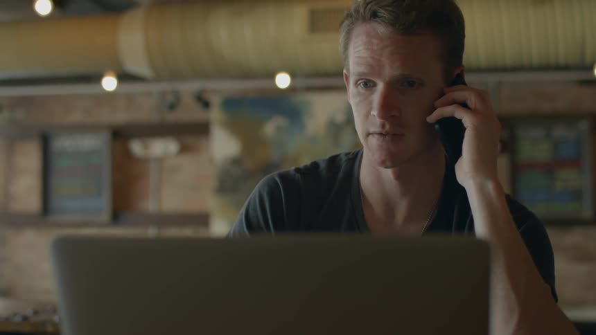 Man talking on his phone while looking at his opened laptop in a business