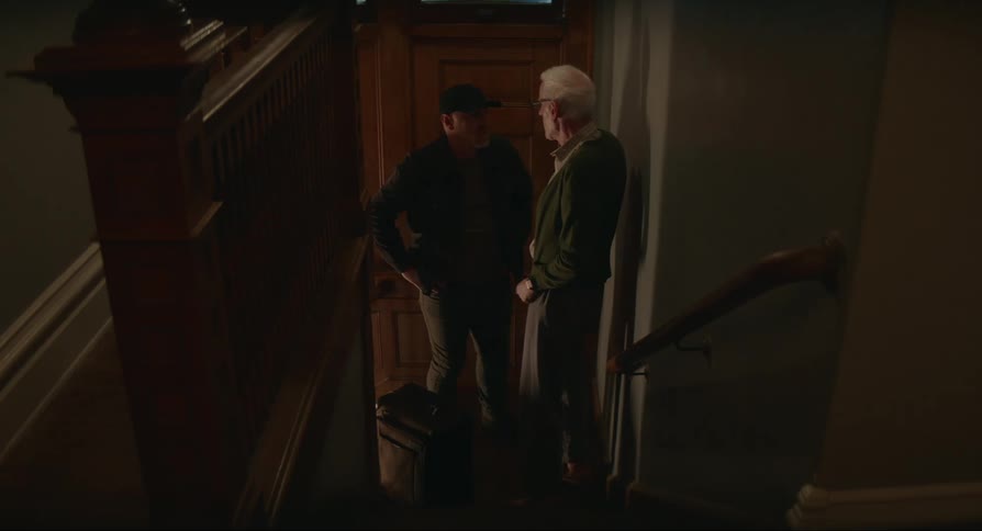 Elderly man wearing glasses greets and hugs a young man wearing a cap and both climb upstairs