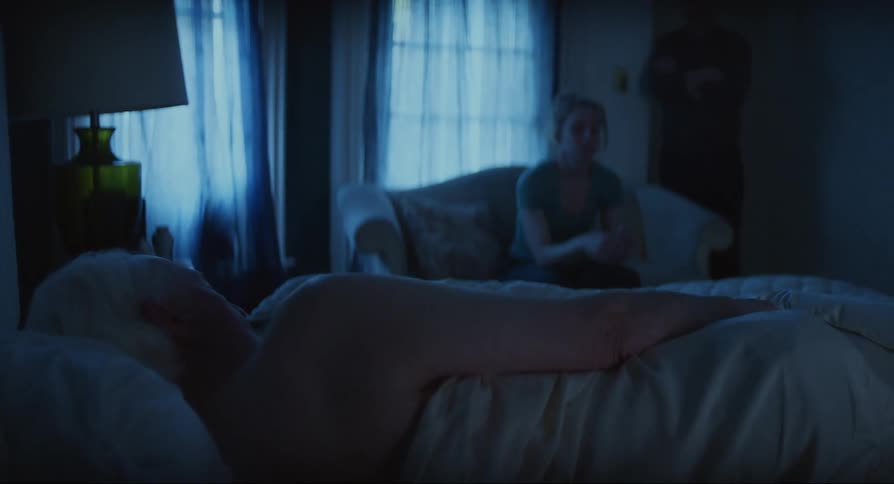 Elderly person lying in bed and another person a woman sitting on the nearby sofa with a sad expression on her face