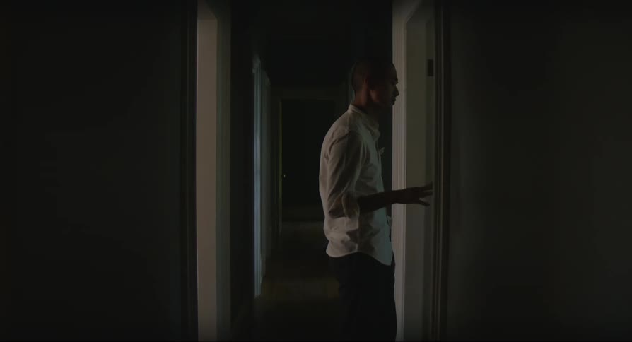 Black man wearing a white shirt indoors walking into a corridor then stopping in front of a door and looking around contemplatively
