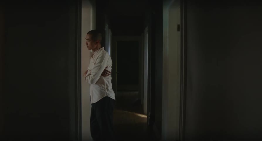 A bald asian man is leaning against a doorway in a hallway