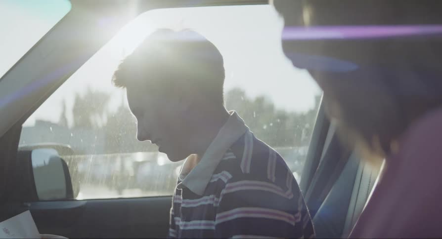 A teenage boy reads a paper and is hugged inside the car by his mother