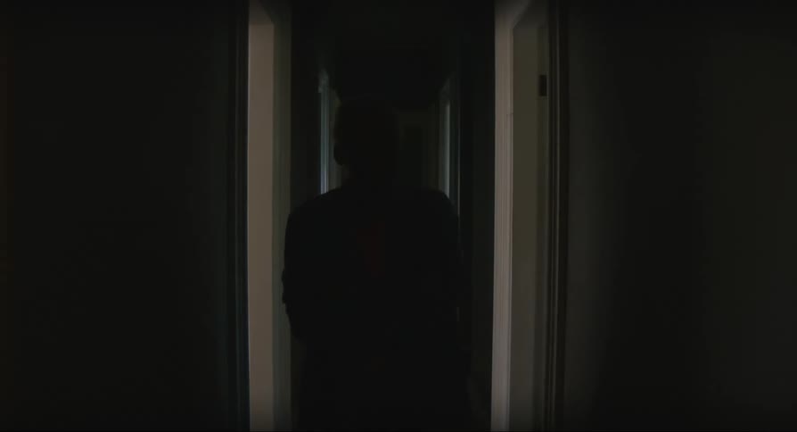 An elderly woman in a robe comes out of a door into a long dark corridor