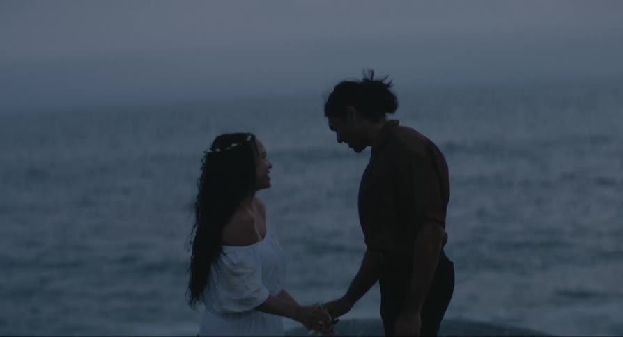 A man and a woman a couple gazing romantically at each other while the woman holds a bottle of wine both beside the sea during a sunset