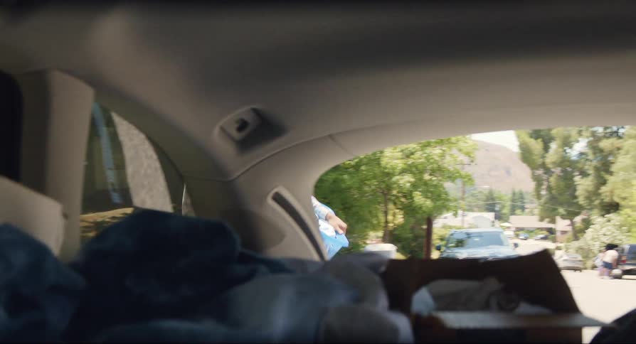 Black woman hugging black teenager boy outdoors in front of a house before he carries a cardboard box and a plastic basket and loads them into the trunk of a car during day time