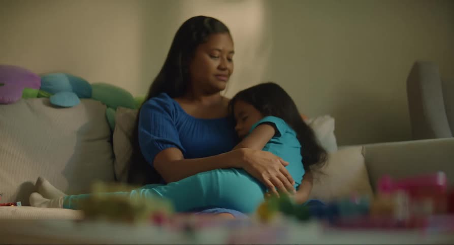 Mother sitting on the couch holding her daughter in her arms as she sleeps with a table in front of her displaying toys