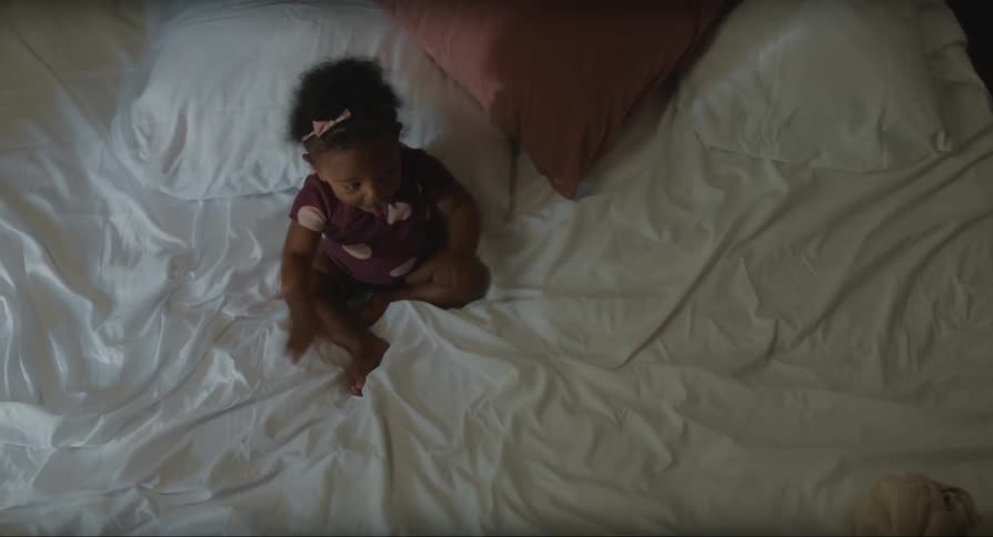 A baby girl in a polka dot outfit sitting on a bed slapping her legs before crawling away