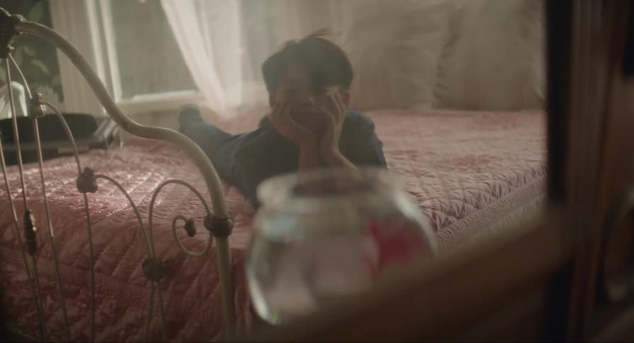 Mirror reflection of an asian boy lying down on a pink bed on a bedroom indoors looking at a fish bowl