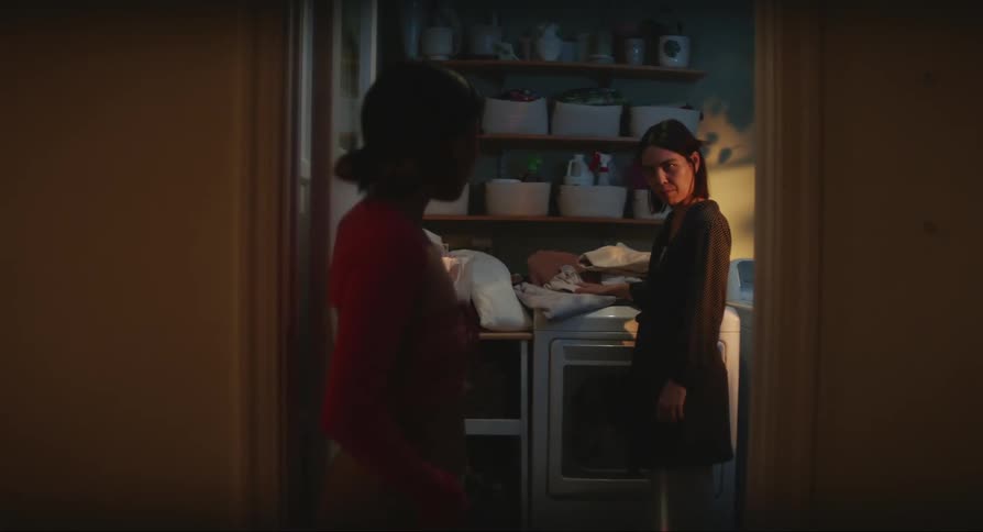 A mother folding laundry as her daughter walks by and they have a conversation while the girl holds her phone