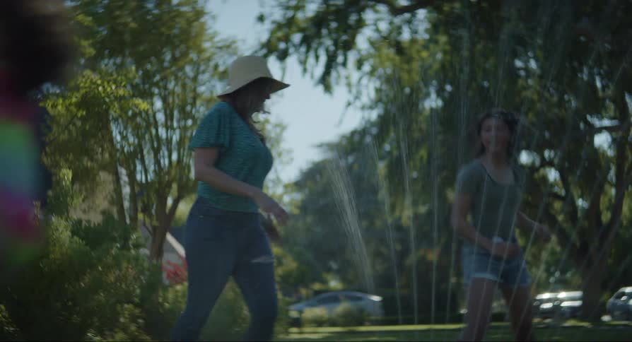 Woman and girls jump over lawn sprinkler in the yard