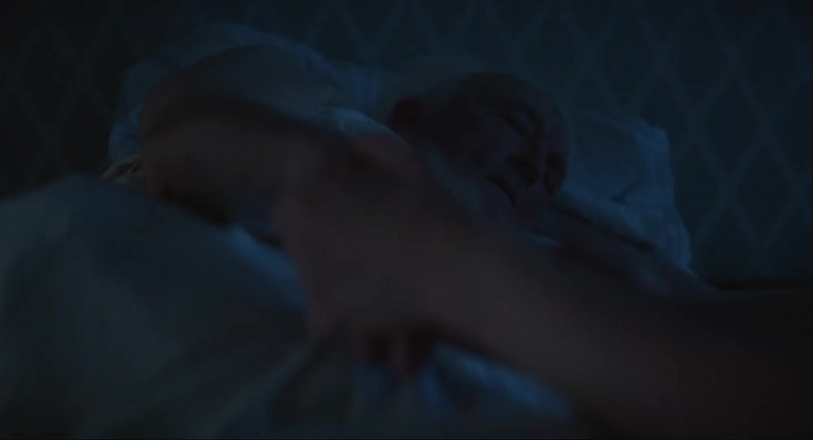 An old man is sleeping lying on a bed and a person is holding his hands