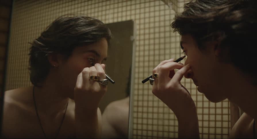 Close up of a male putting black eye makeup on his face in a mirror in a dimly lit room