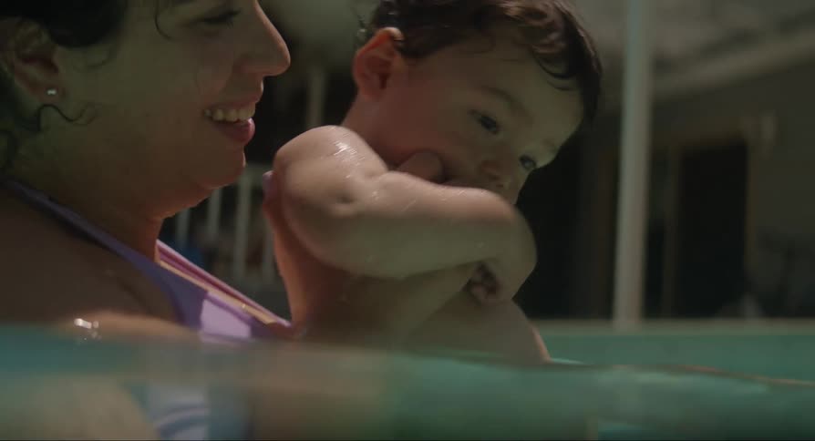 A toddler by being held by his mother in a swimming pool next to the father on a bright sunny day
