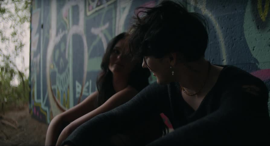 A teenage girl and boy sitting against a graffiti wall as they talk