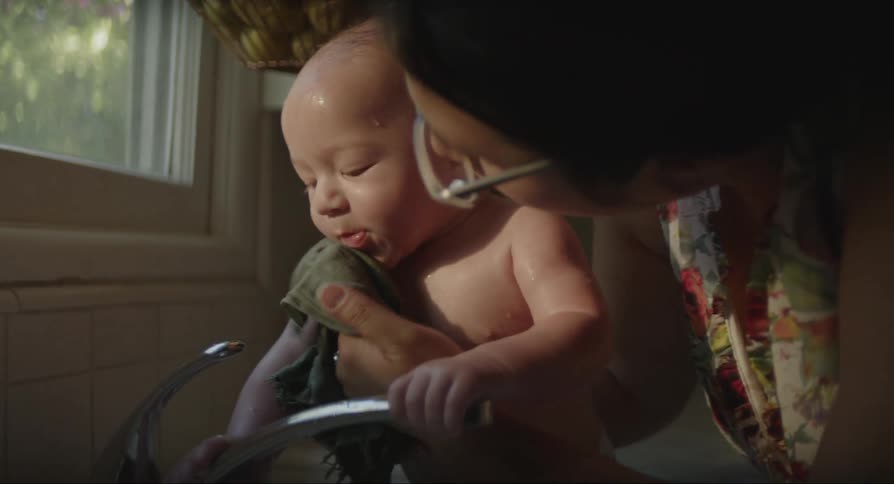 A baby getting bathed in a kitchen sink by his mother on a bright day as the sun shines through the window