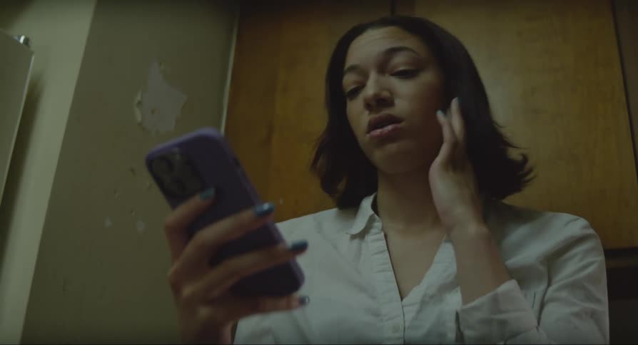 A woman is sitting down holding her cell phone and she rubs her face with her hand