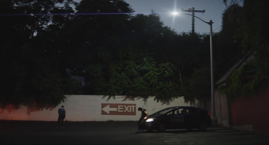 Two people outdoors at night with one of them skateboarding and the other observing while leaning against a car with its headlights on