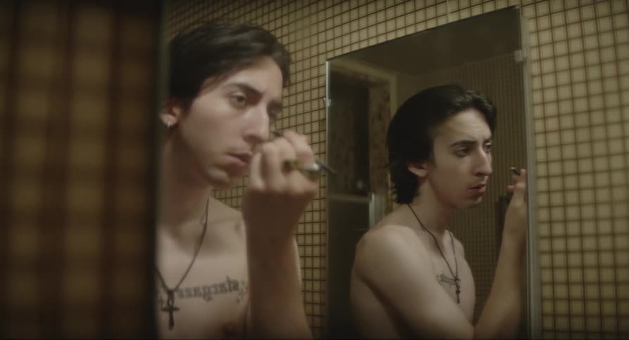 A shirtless young man in a bathroom putting on eye makeup while looking into a mirror
