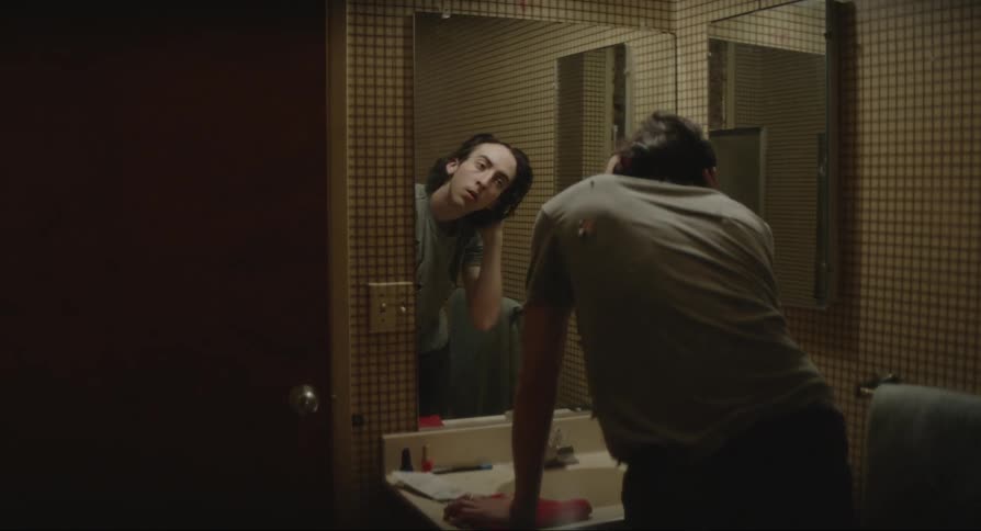 A boy standing in front of a mirror in a bathroom as he combs his hair back looking at his reflection
