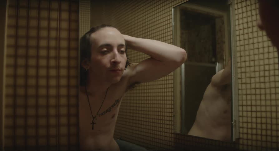 A boy standing in a mirror with no shirt on as he runs his hand over his face