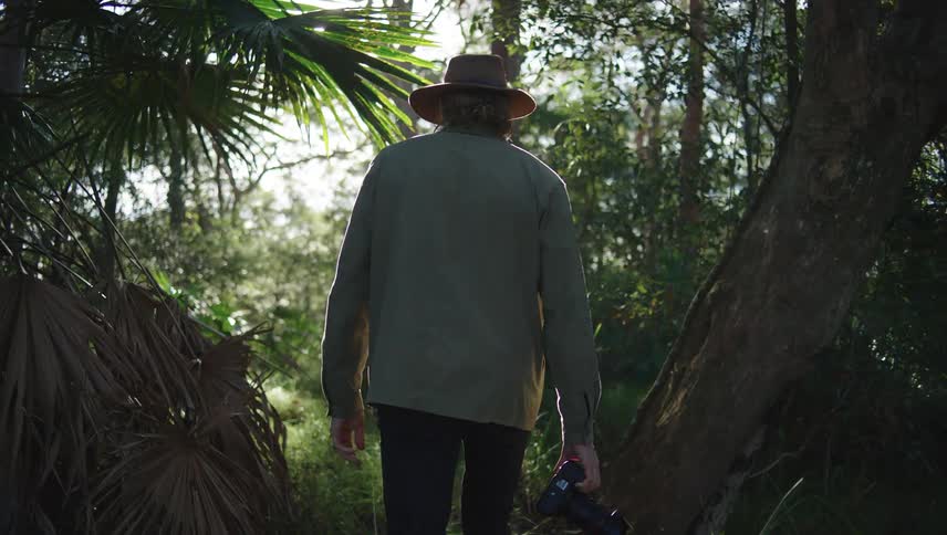 Photographer walking through a forest with his back turned holding his professional camera in one hand