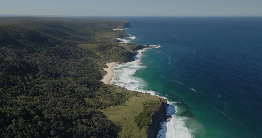 Drone view of ocean waters crashing up against a green coastline full of trees on a bright sunny day