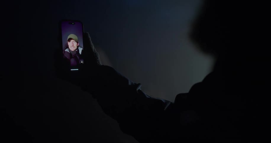 A silhouetted person in the dark at night is talking by video to a man on a smart phone