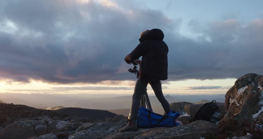 A man is standing on high ground with a camera on a tripod as he turns and steps away