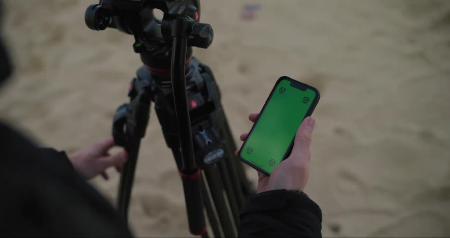 Human hands moving the screen of a cell phone and next to it a tripod for a camera