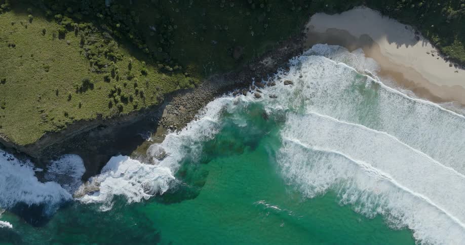 Drone view of ocean waves crashing against the sand near a grassy land on a bright sunny day