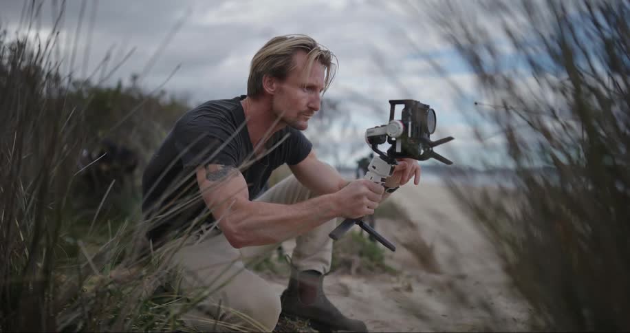 A blond man with a tattoo is bloated in the sand holding a digital camera