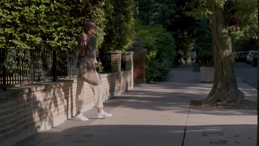 A man sits on a small wall and then takes his bag and leaves on the sidewalk
