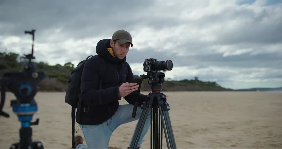 A man wearing a cap black coat jeans and a backpack adjusts his camera on a tripod at the beach