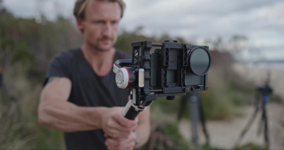 A man standing before tall grass on a beach sets up a camera pointed toward the ocean