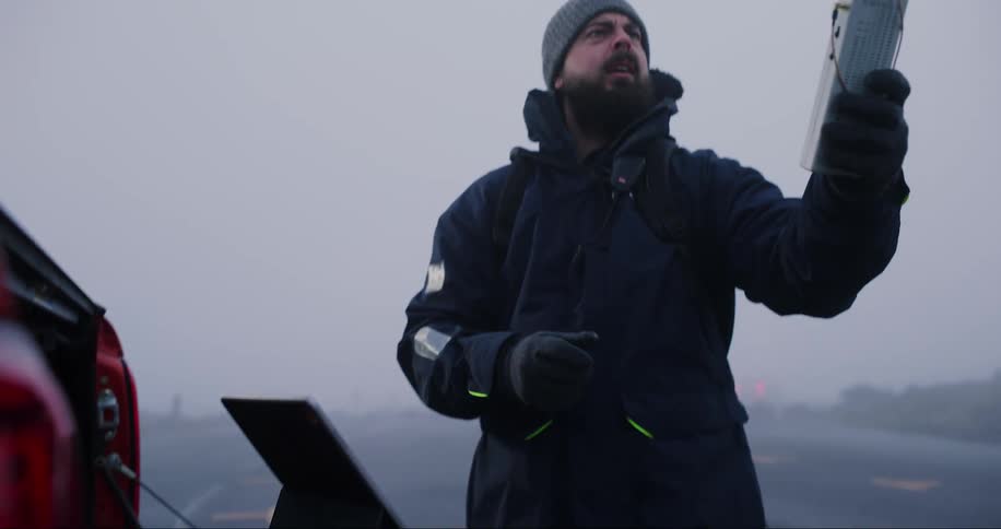 A man in cold weather clothes working on a laptop and pointing a device in his hand upwards
