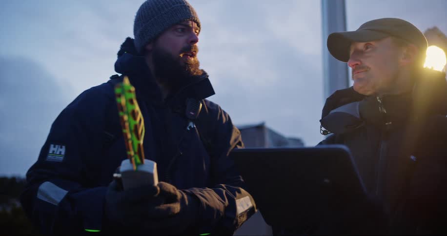 Two men wearing winter coats one holding a tablet the other holding a piece of equipment while they stand outside