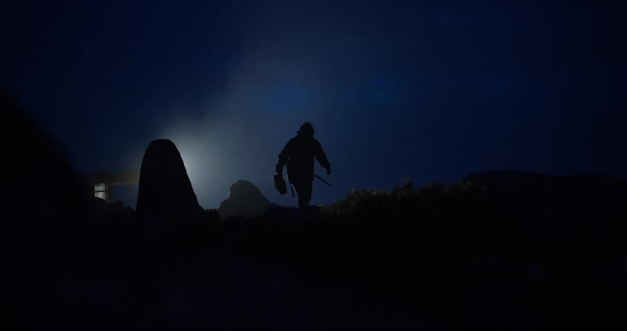 Silhouette of a man walking across dark covered land at night as light shines from behind a rock formation