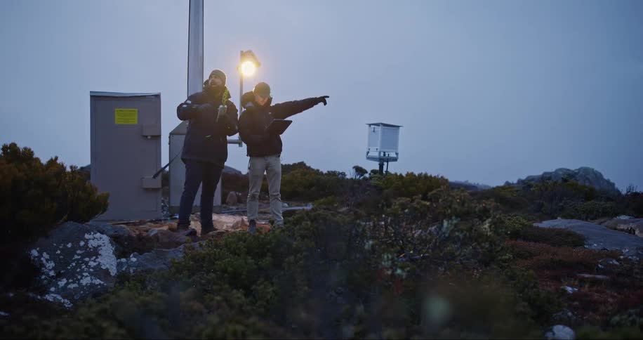 Two men are standing along the coast near a utility tower with a tablet computer and an electronic device as the point and talk