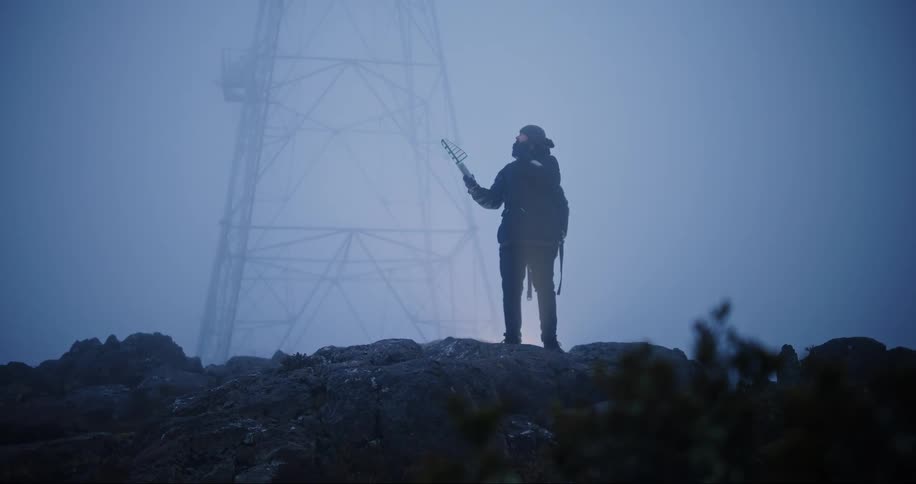 A man with a device in his hand walks up a hill and lifts his hand with the device and then leaves
