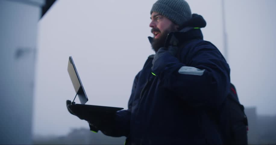 A man wearing a winter coat and hat holds a laptop computer as he looks at a utility box and speaks into a microphone on his coat and walks away
