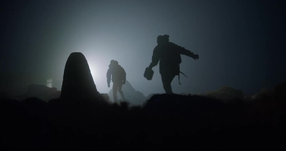 Two men walking across rough terrain at night toward a light wearing coats and carrying equipment