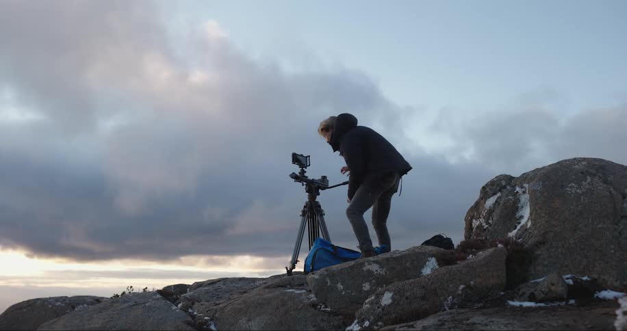 A man standing on a rocky cliff in the day sets up a camera