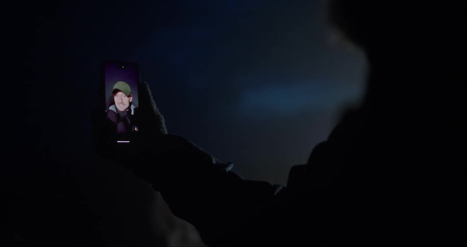 A person holding a cellphone with a video of a man on the screen at night in a very dimly lit area