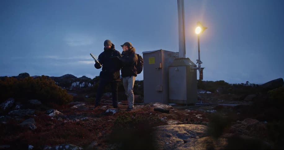 Two men holding a laptop and an antenna conversing on top of a mountain at night above the cloud level