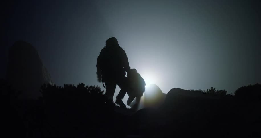 Silhouettes of two men walking up a hill and one man is carrying a briefcase as a light shines very brightly in the distance
