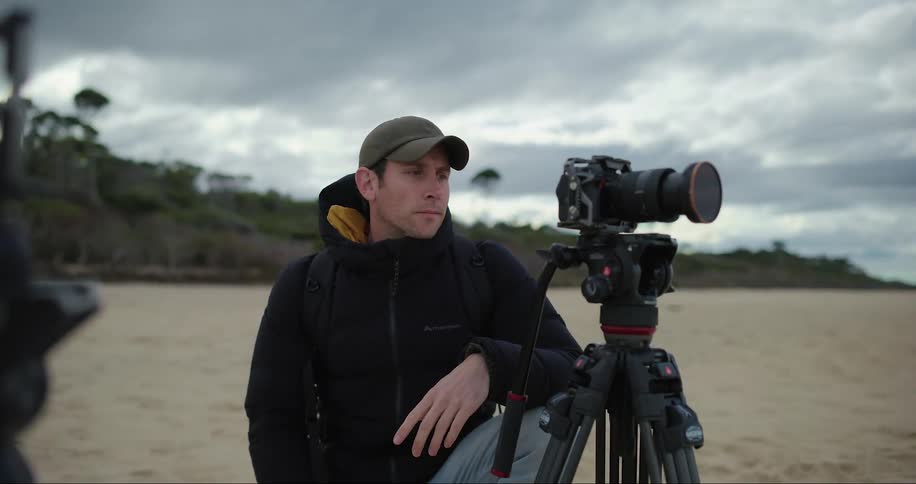 A man in a hat and coat tied up in the sand taking pictures with a professional camera on a tripod