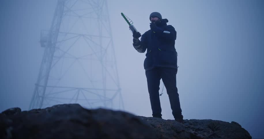Man holding a tracker next to an antenna on top of rocks then communicating with someone through a communication device