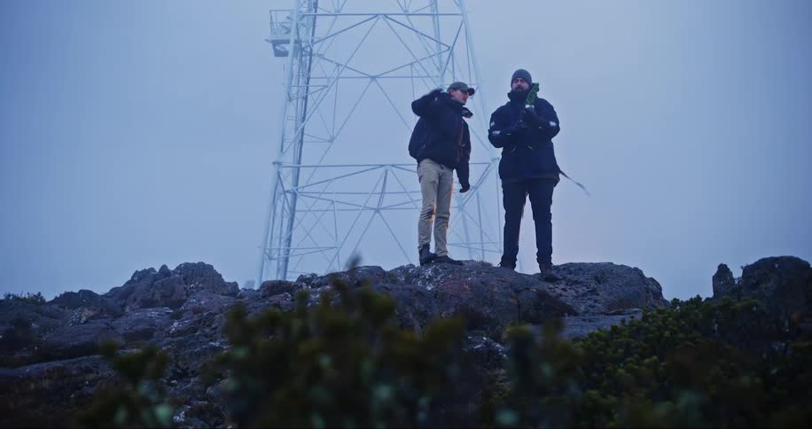 Two men wearing coats and hats walking on a rocky ridge holding equipment in front of a tower