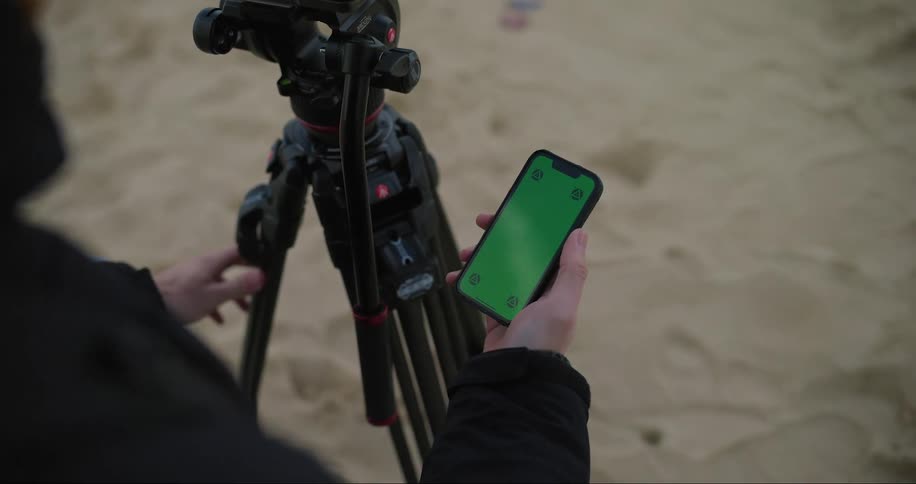 A man is standing in the sand next to a tripod as he holds a cell phone with a green screen